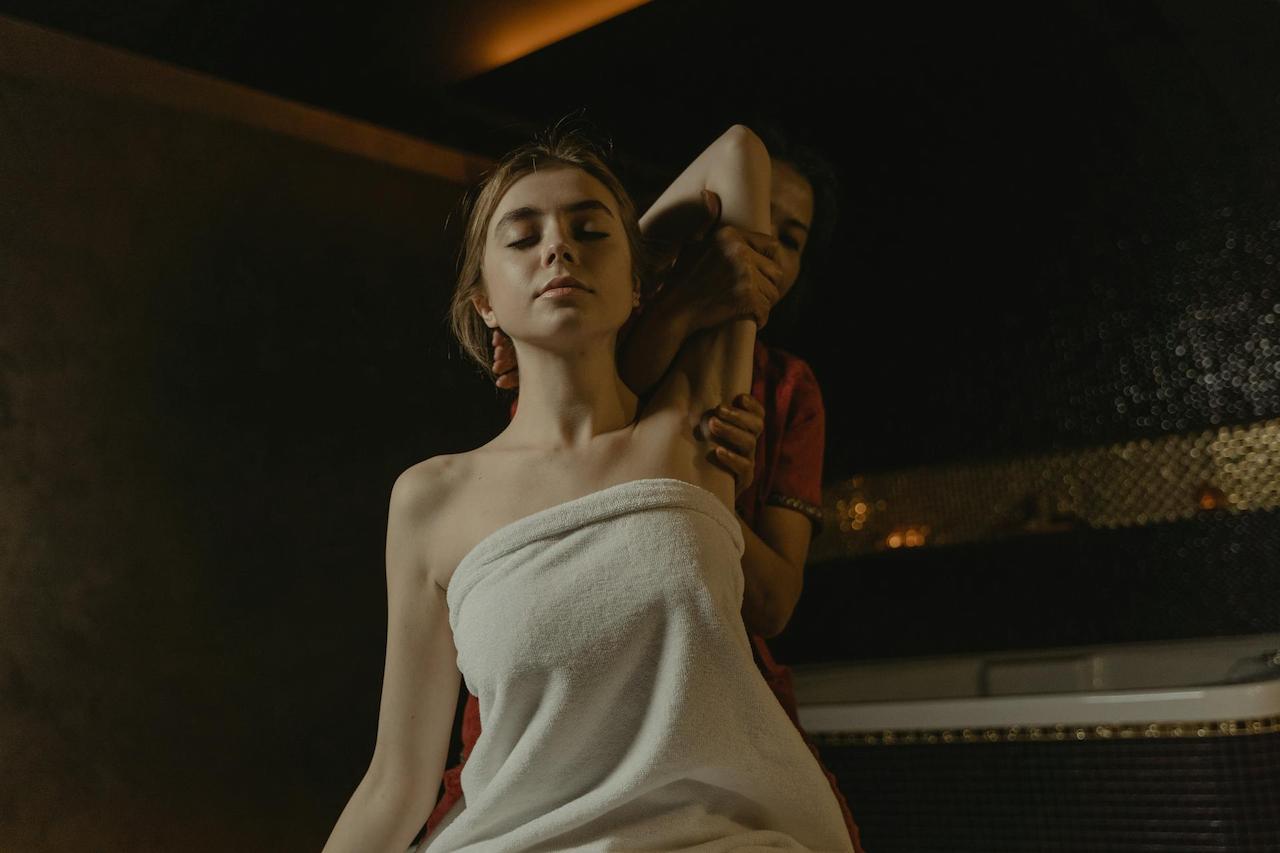 The masseuse is lifting the blonde lady's left arm during the Thai massage session.
