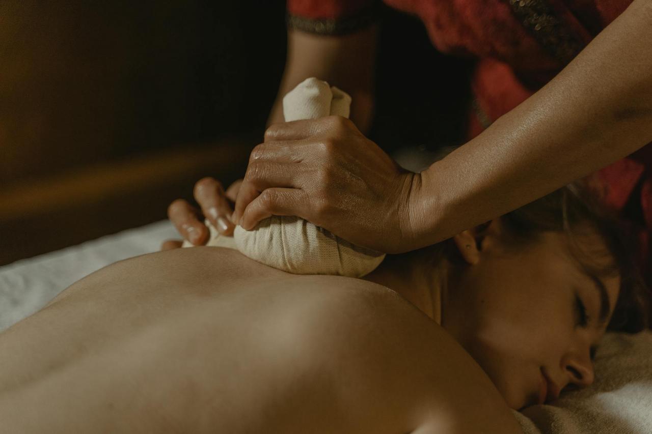 The therapist applies steamed herbal compresses filled with fragrant herbs to the upper back area of the client during the Thai Herbal Massage session.