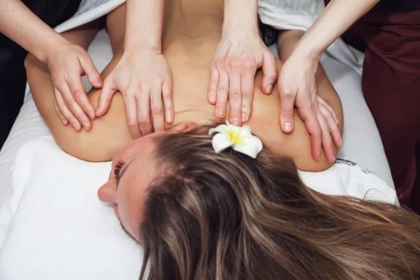 Two skilled therapists are working simultaneously to provide a synchronized and deeply relaxing four-hand massage on the lady's back.