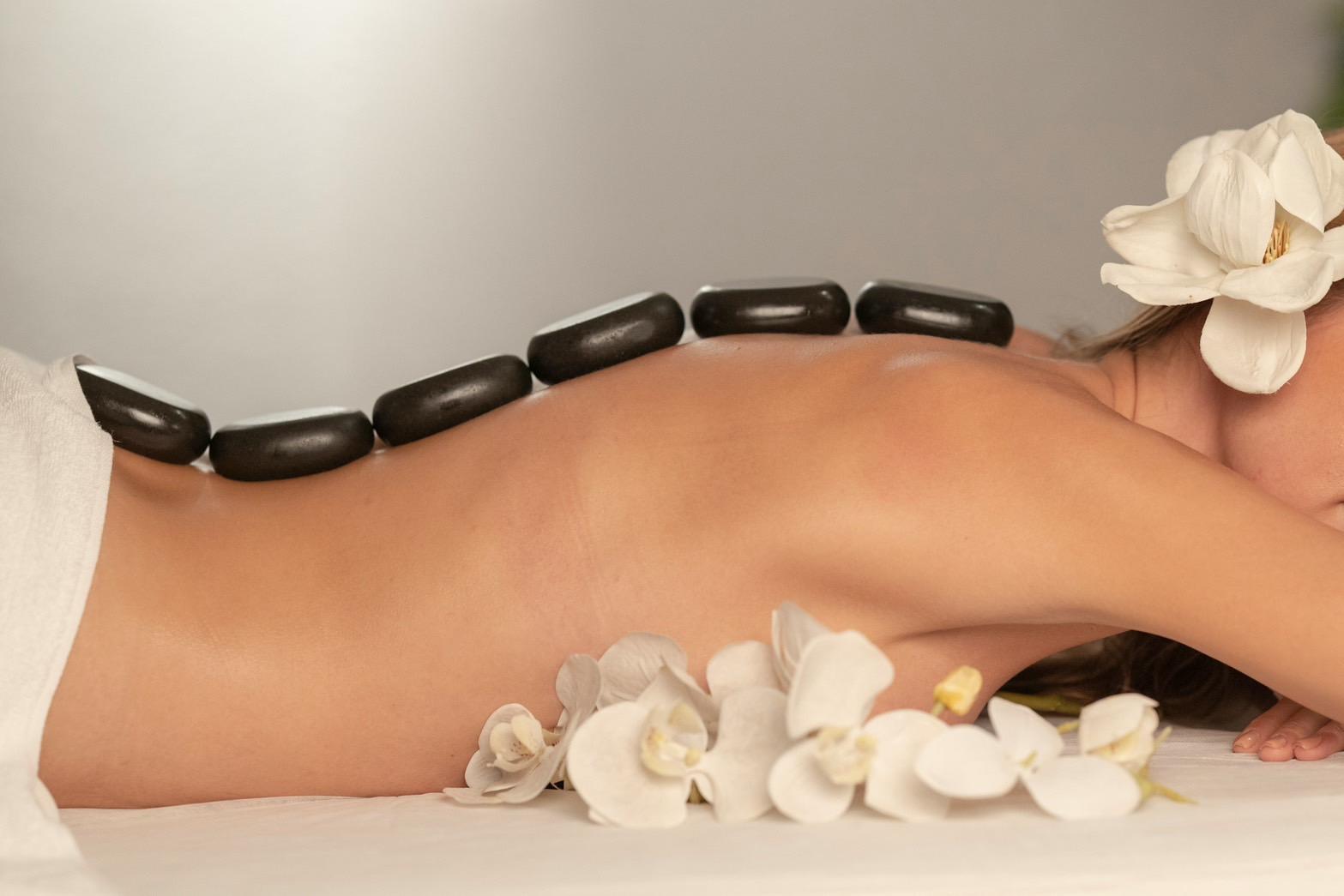The woman lies down on her front. The therapist is putting hot stones on her back during the hot stone massage session. The mattress is full of white flowers.